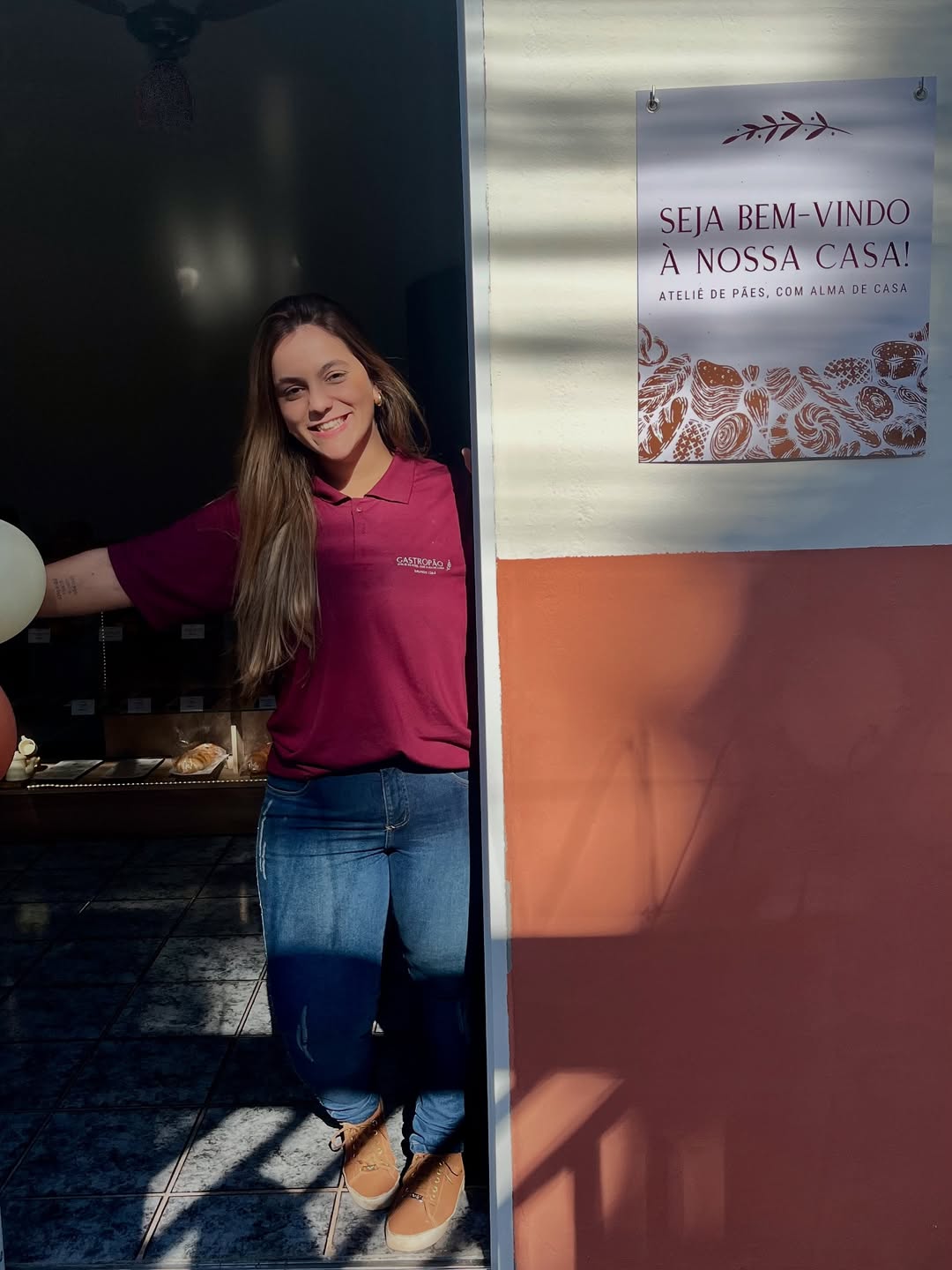Bia Sant'Anna, fundadora do Gastropão, na entrada da loja com o cartaz Seja Bem-Vindo à Nossa Casa